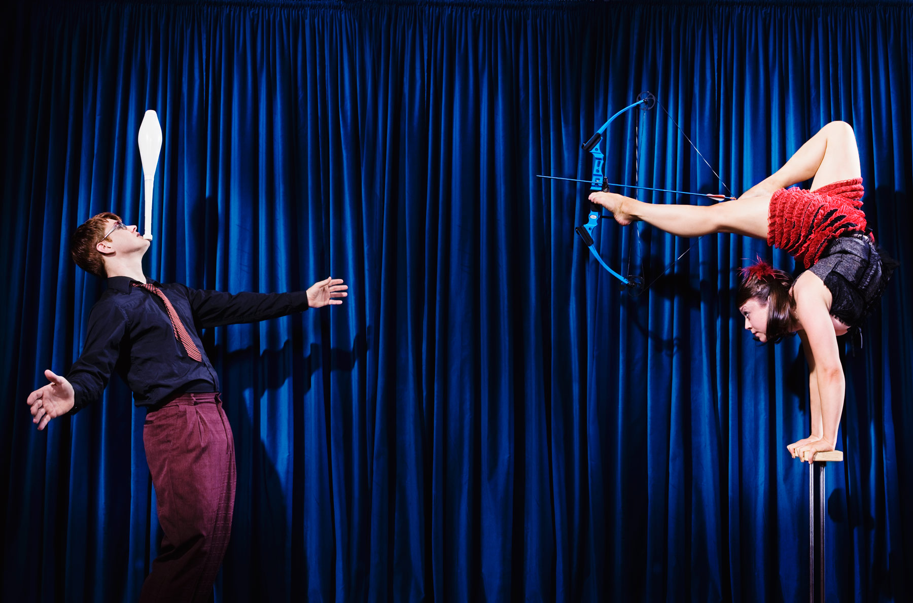 Two performers doing a trick with the man balancing a pinball on his chin and the woman holding a bow and arrow with her feet while balancing on her hands.