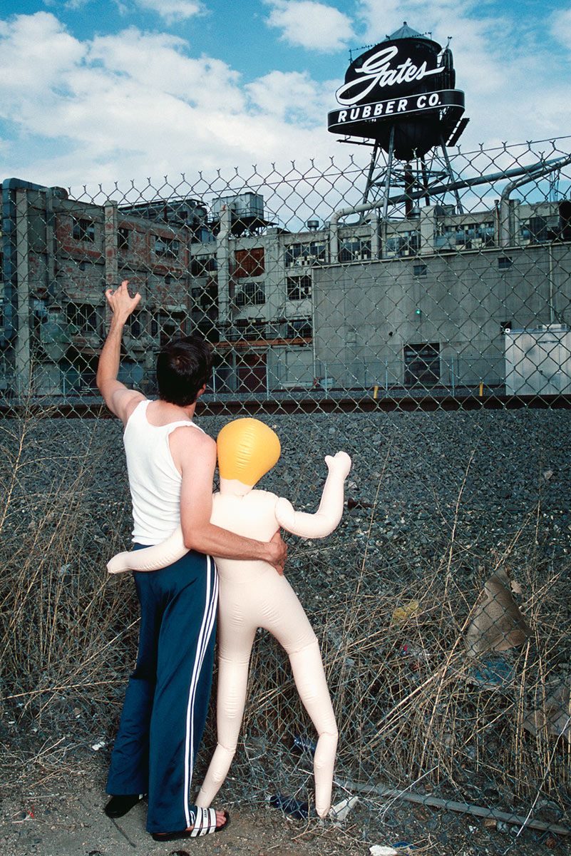 A man and blow up doll embracing against chain link fence with view of  Gates Rubber Factory.