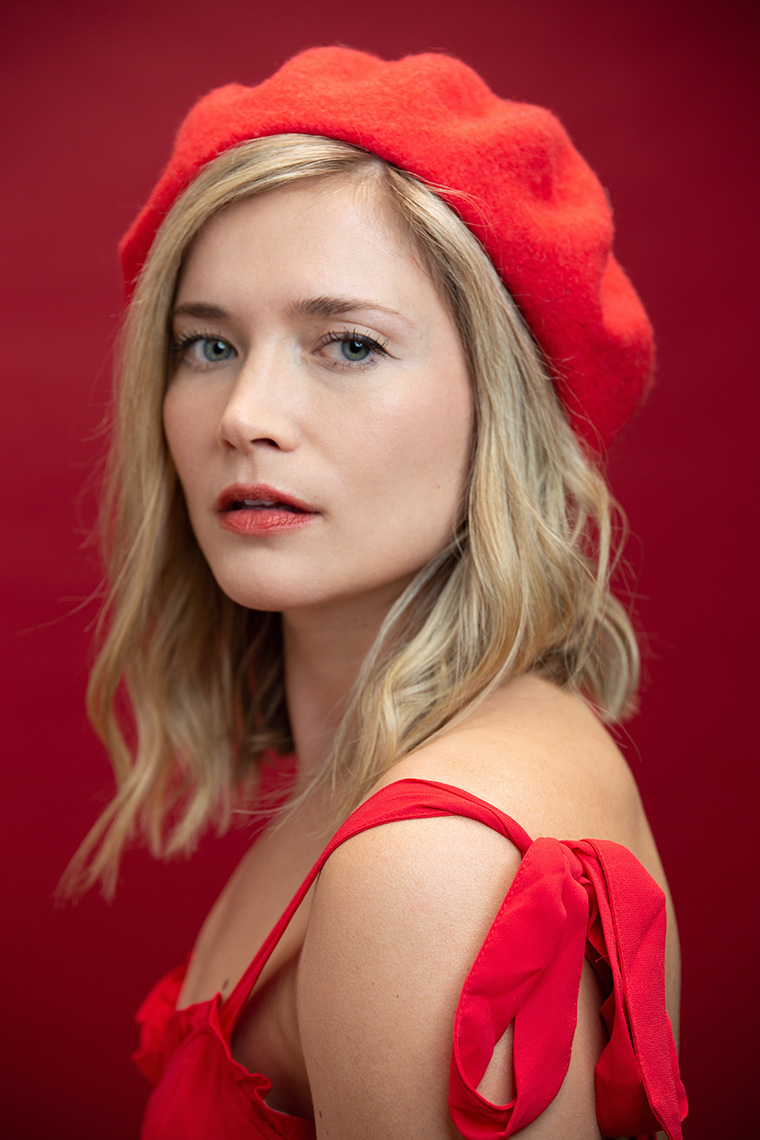 Woman with blonde hair  wearing red dress, red hat in front of a red background.