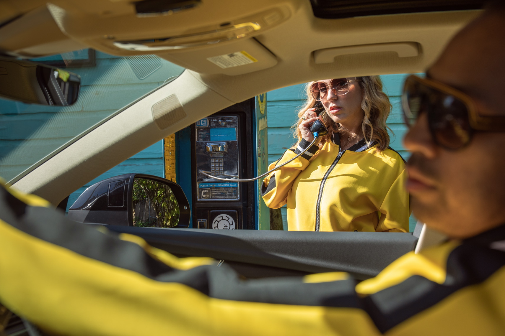 Woman at phone booth, man in car, with both wearing sunglasses and yellow track suits.
