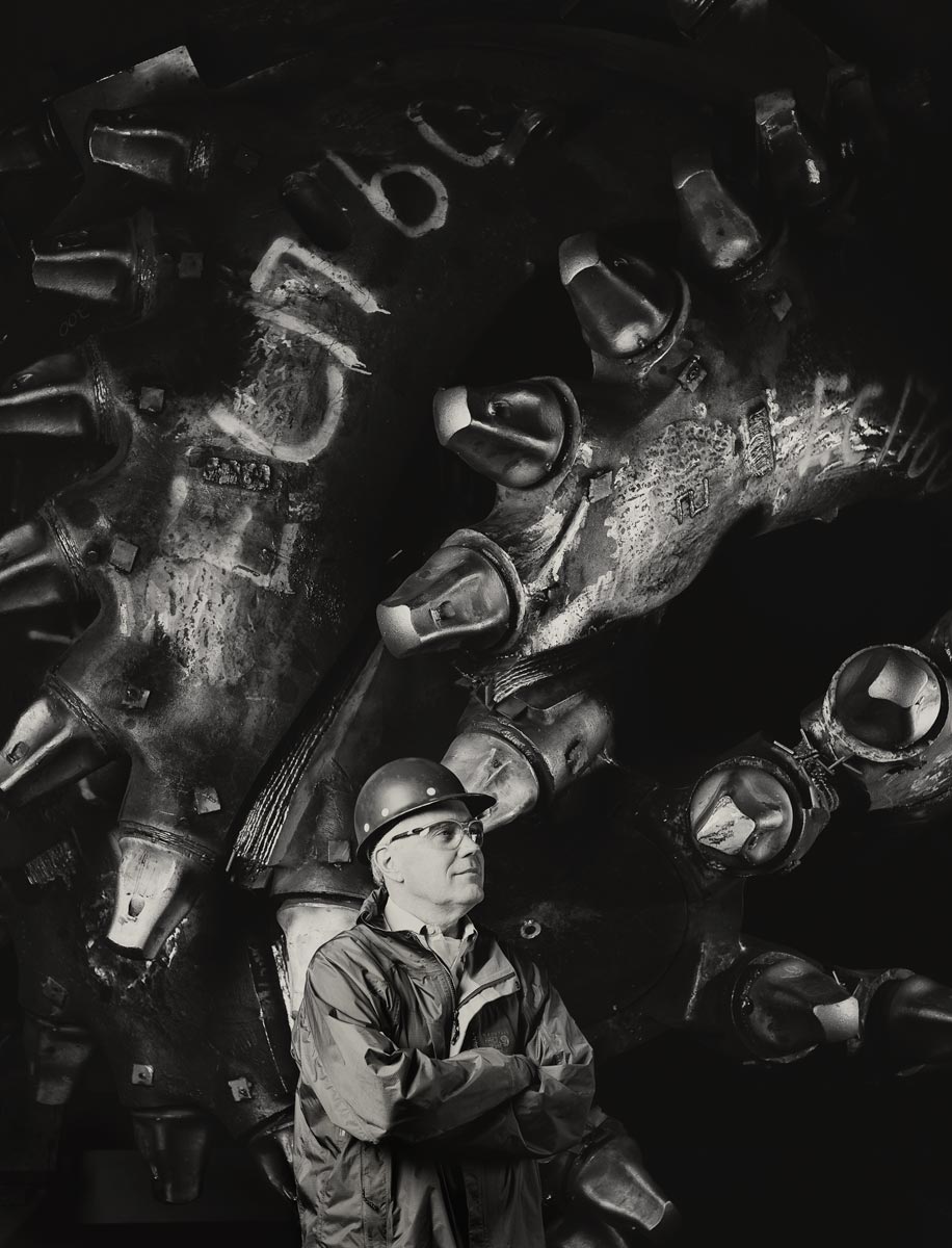 Black and white portrait of man in front of massive dredging drill head.