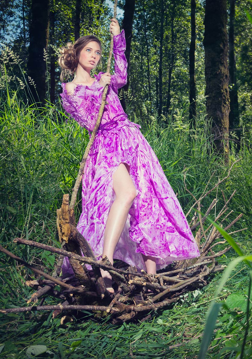 A woman in a stick boat wearing a handmade purple dress located in a field of grass and trees.
