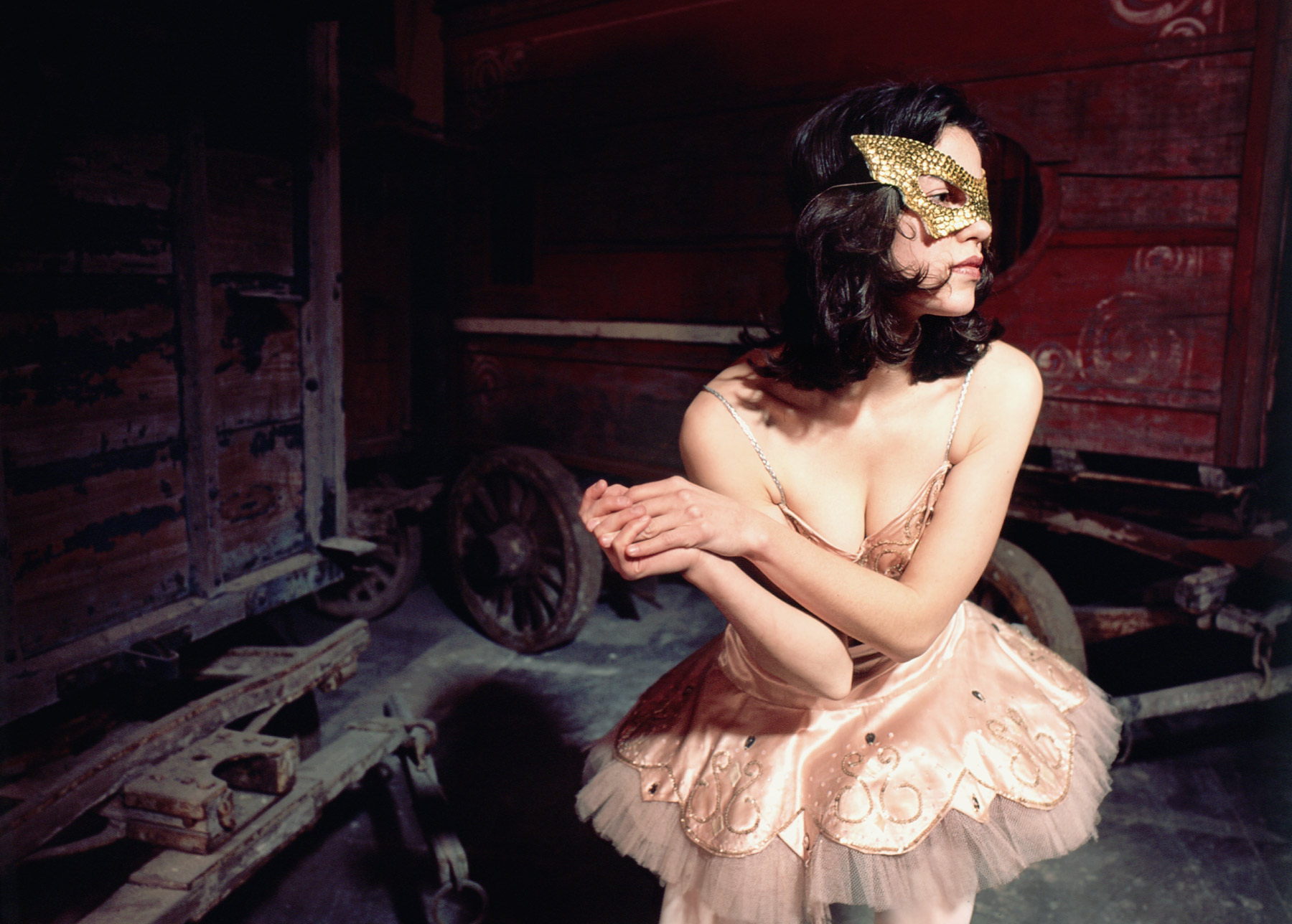 Woman wearing a ballerina costume and gold mask surrounded by vintage circus wagons.
