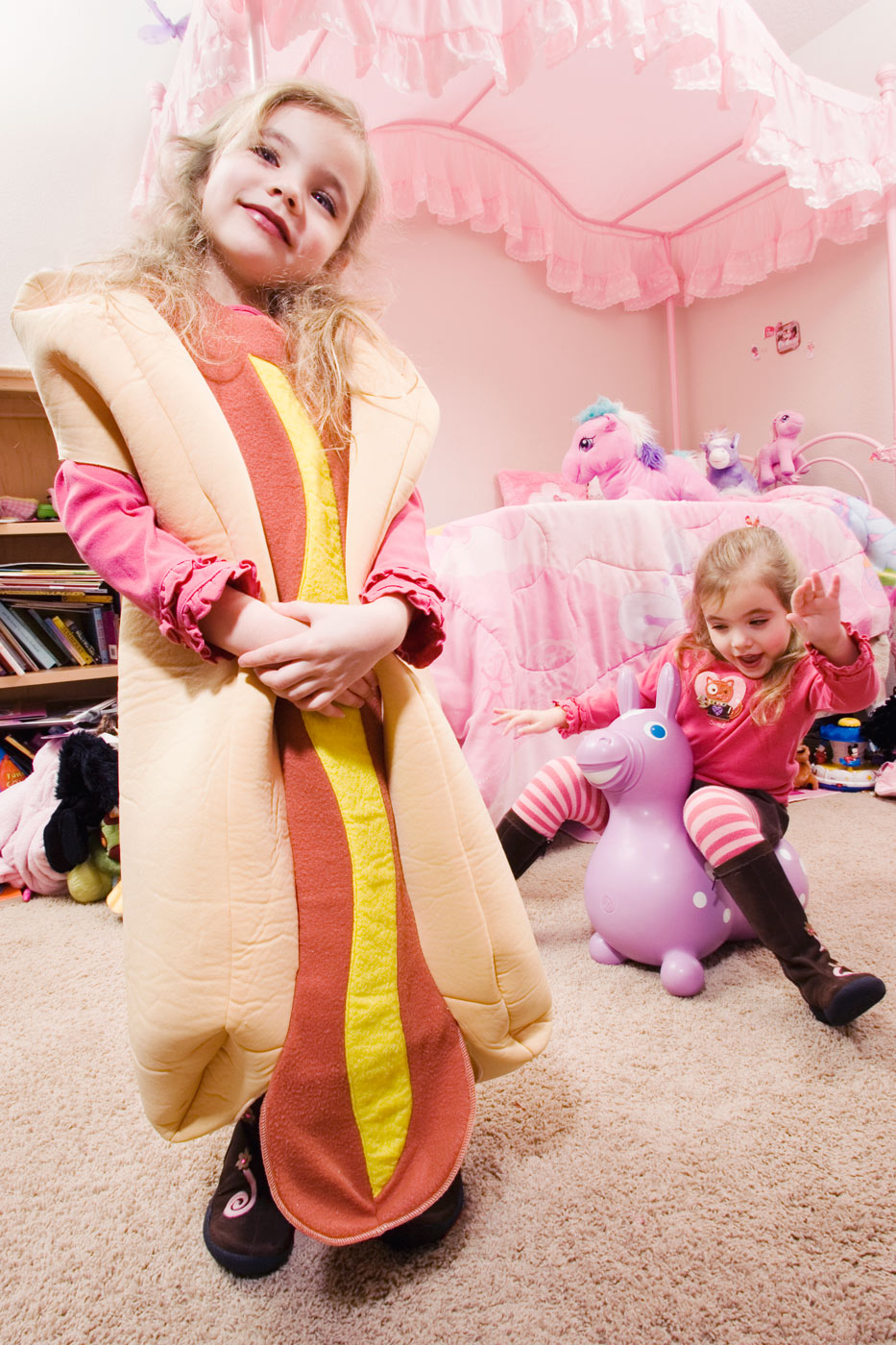 A girl dressed up as an Oscar Mayer hotdog with sister in background  in a pink room with her sister
